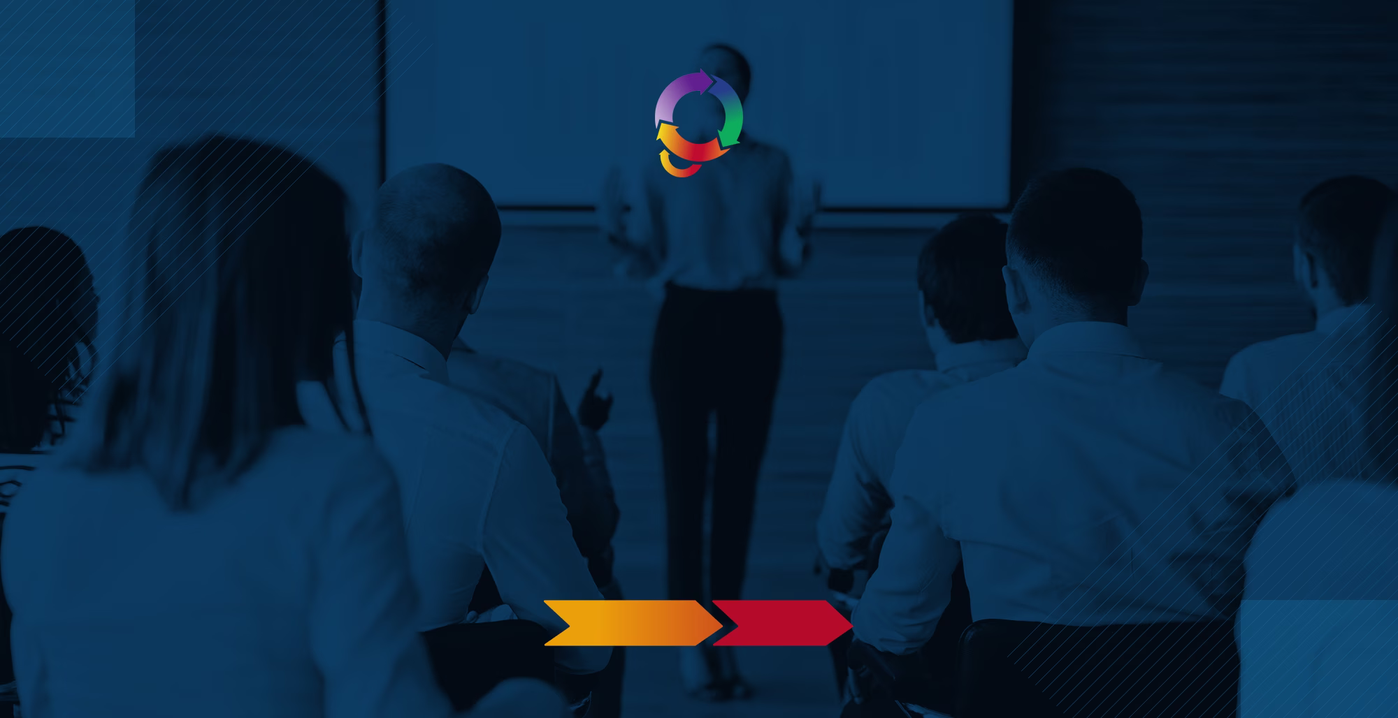 Presenter speaking to an audience in a dimly lit conference room with a multicolor circular logo centered above the stage
